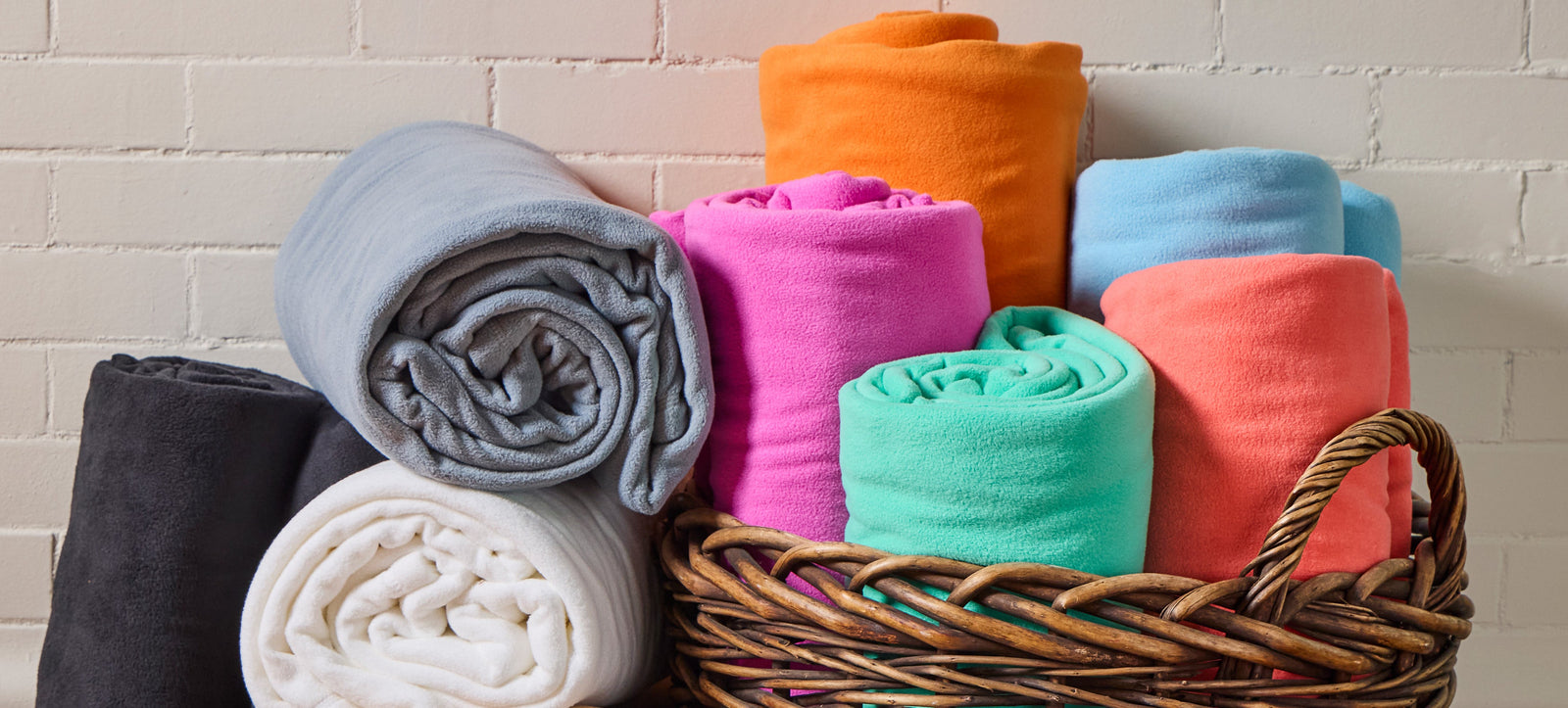 American Blanket Company - Peaceful Touch Fleece Blankets - Pictured is multicolor fleece blankets rolled up and placed in a wicker basket in front of a white brick wall