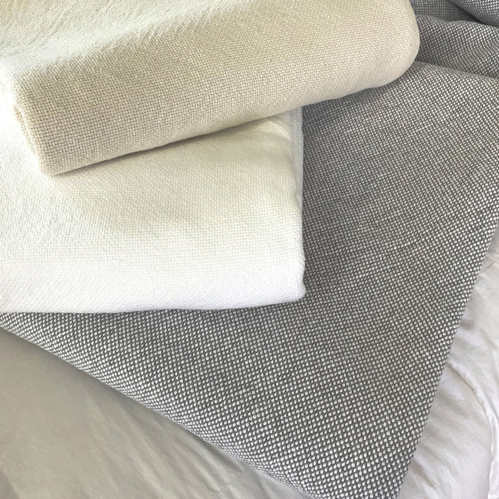 American Blanket Company - 100% Cotton Blanket, Cotton Sweater Weave Blanket - Folded Cotton blankets ontop of a cotton sheet, showing the cream, white, and gray white colored sweater weave blankets.