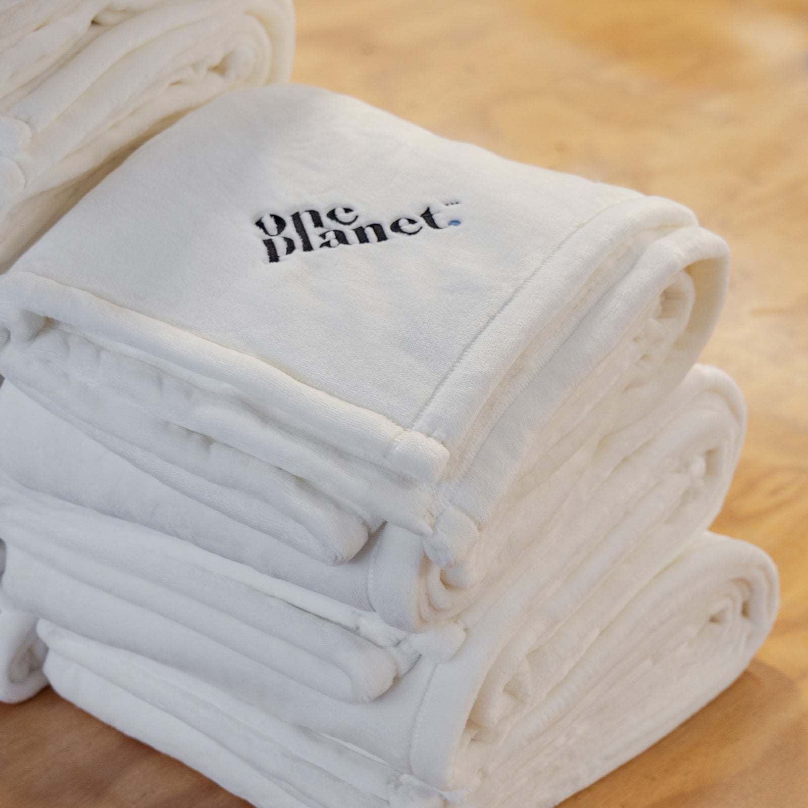 American Blanket Company - Luster Loft Corporate Orders - Stack of white  fleece blankets with 'one planet' logo on a wooden surface