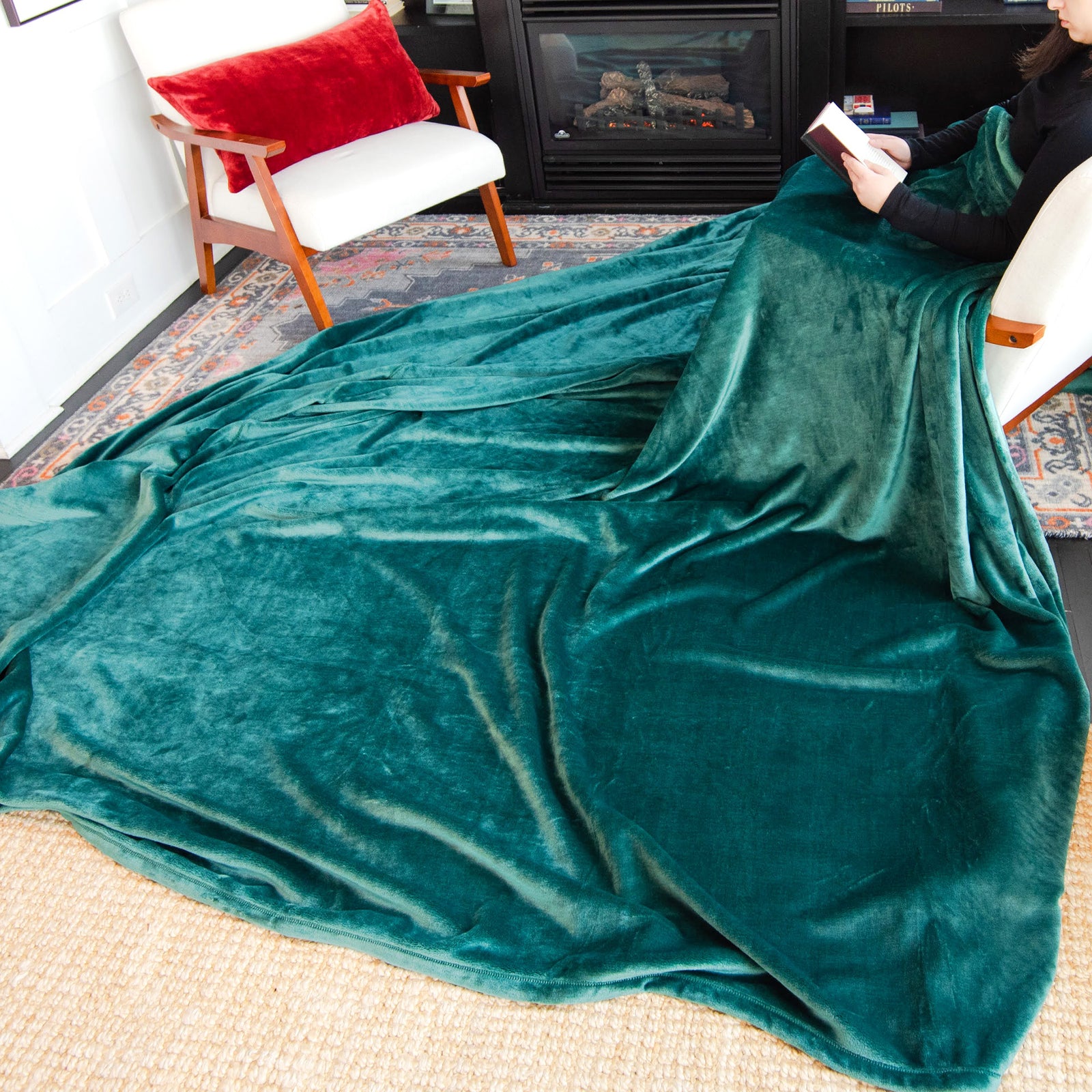 American Blanket Company - Luster Loft Oversized Blankets - Pictured young woman reading in front of a fireplace and have evergreen luster loft fleece blanket cascading over her lap and over the floor by 2-3 feet