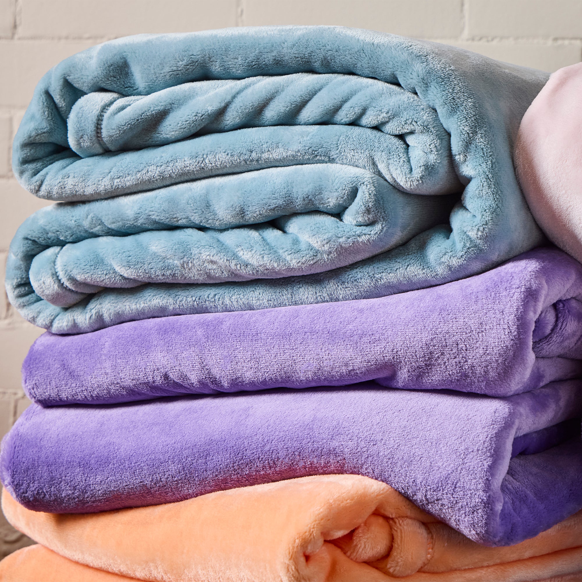 American Blanket Company - Luster Loft Light Weight Fleece Blankets - stack of folded fleece blankets in blue, purple, and orange on a wooden surface with a brick wall background.
