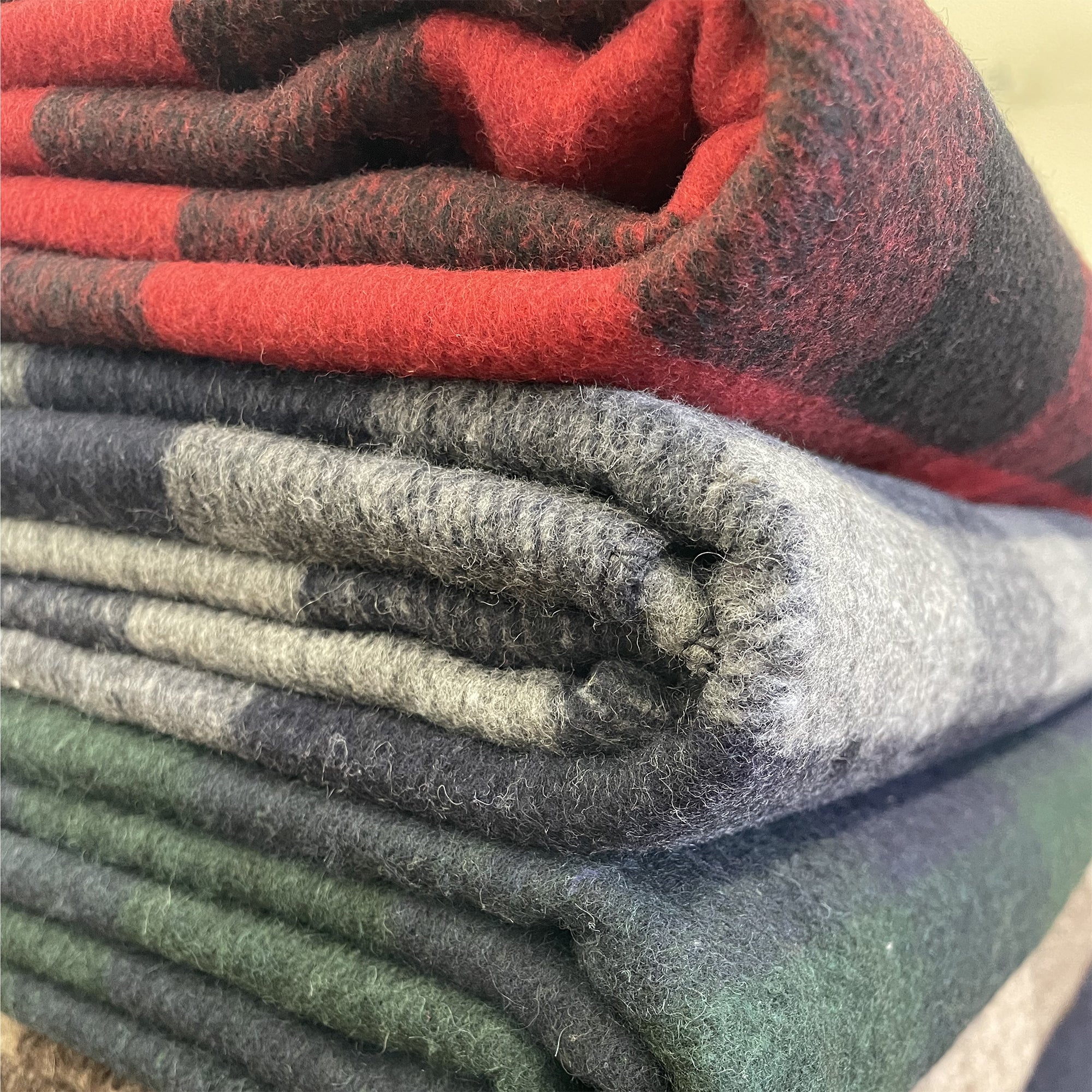 American Blanket Company - Recycled Wool Blankets - Close up of the the folded Recycled Wool Blanket Line UP On Wood showcasing red black plaid, brown tan plaid, blue gray plaid,green blue plaid.
