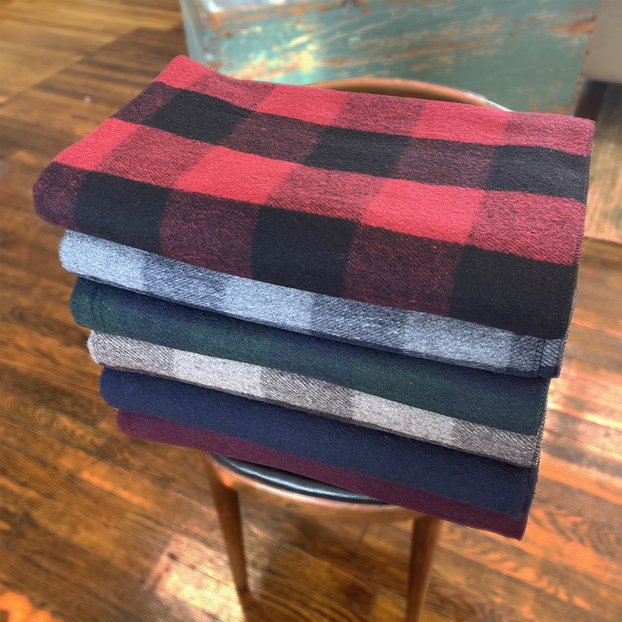 American Blanket Company - Recycled Wool Blankets - Recycled Wool Blanket Line UP On Wood showcasing red black plaid, brown tan plaid, blue gray plaid,green blue plaid folded and stacked on a wooden chair.
