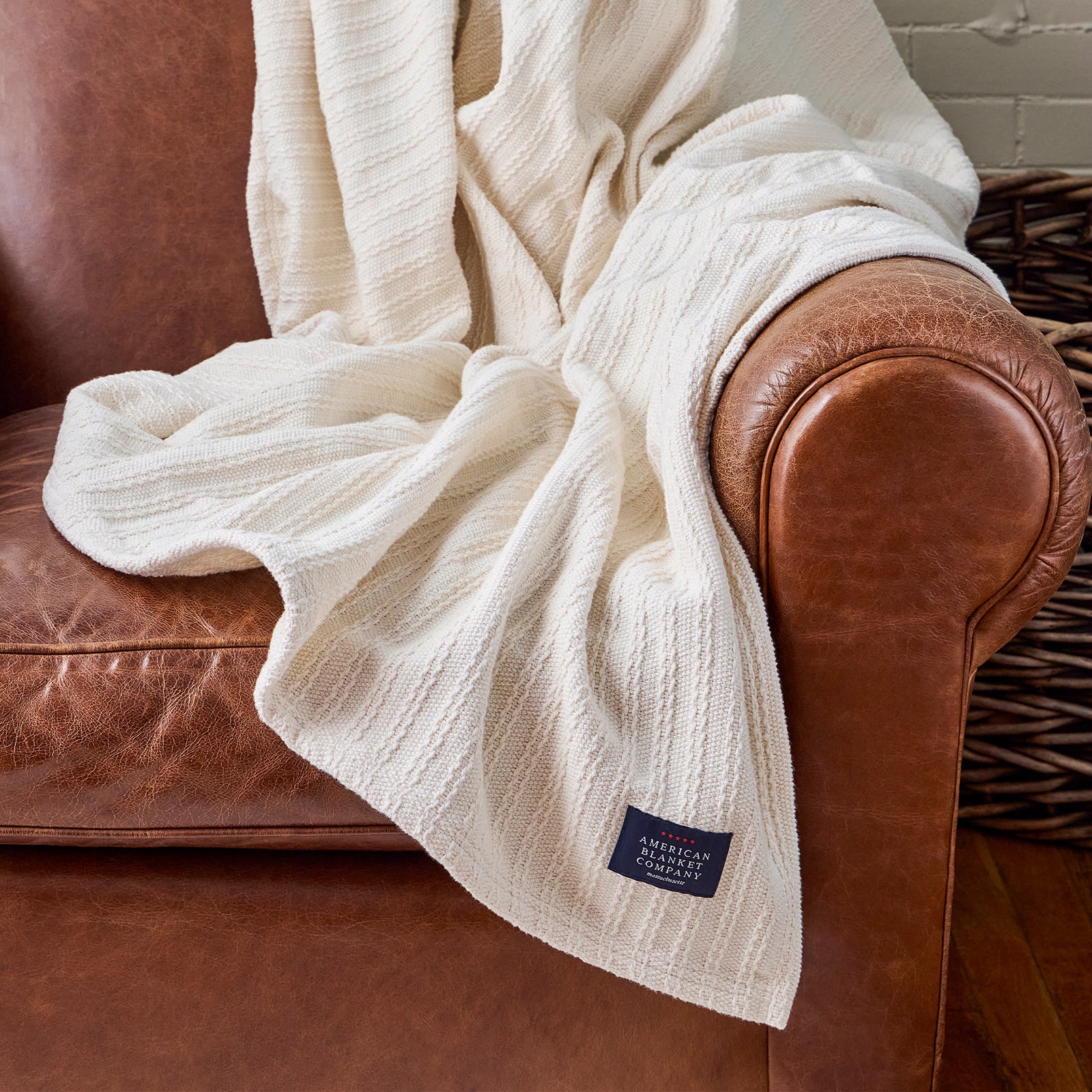 American Blanket Company - Cable Weave Cotton Blanket - Cable Weave Cotton Blanket - White textured blanket draped over a brown leather armchair with a visible brand label.