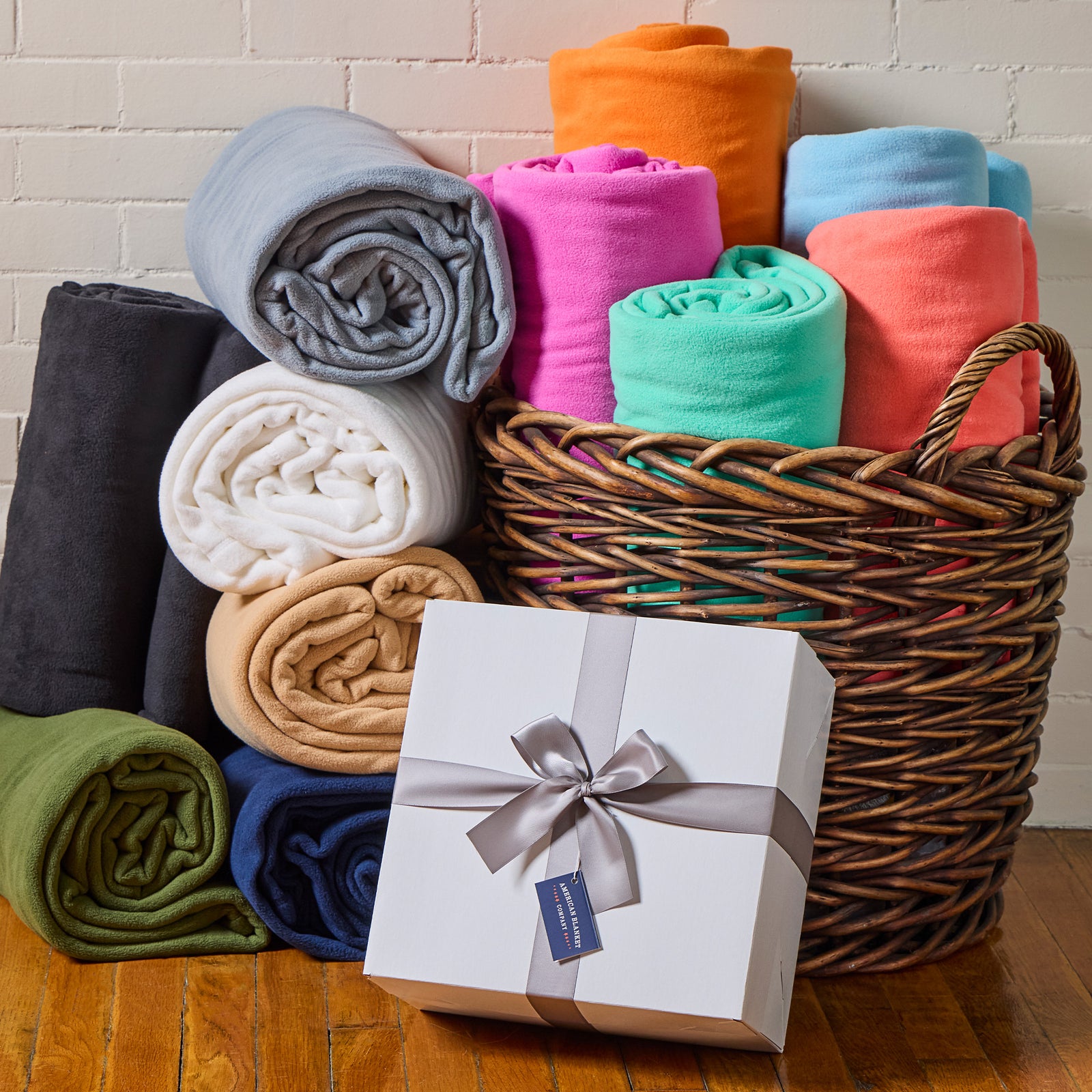 American Blanket Company - Corporate Orders Fleece Blankets - rolls of Colorful fleece blankets in a basket with a gift box and hang tag reading "American Blanket Company" on a wooden floor in front of a white brick background.