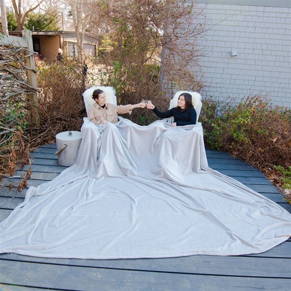 American Blanket Company - Luster Loft Fleece Oversized Blankets - Pictured are two woman toasting outside on a blue wooden deck, both covered by a large luster loft blanket cascading over both of the laps over the blue deck.