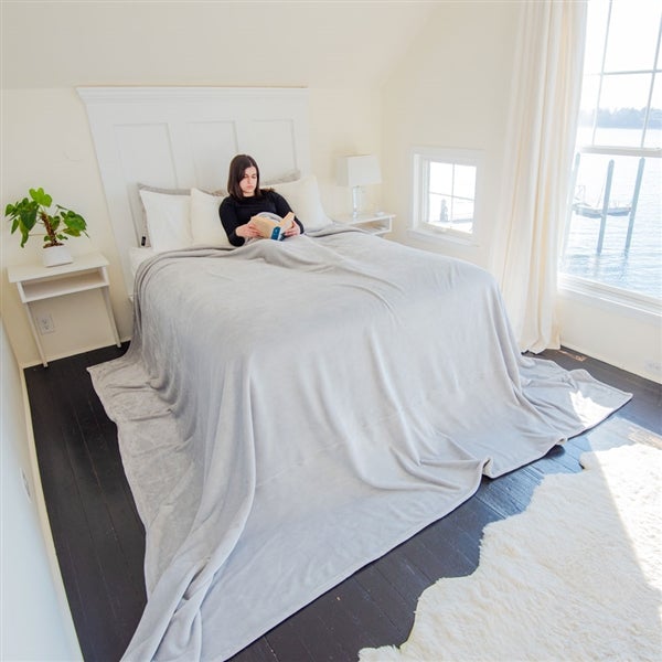 American Blanket Company - Luster Loft Fleece Oversized Blankets - pictured young woman on a large bed draped in a light gray luster loft oversized blanket that falls over the bed and past it onto the floor.