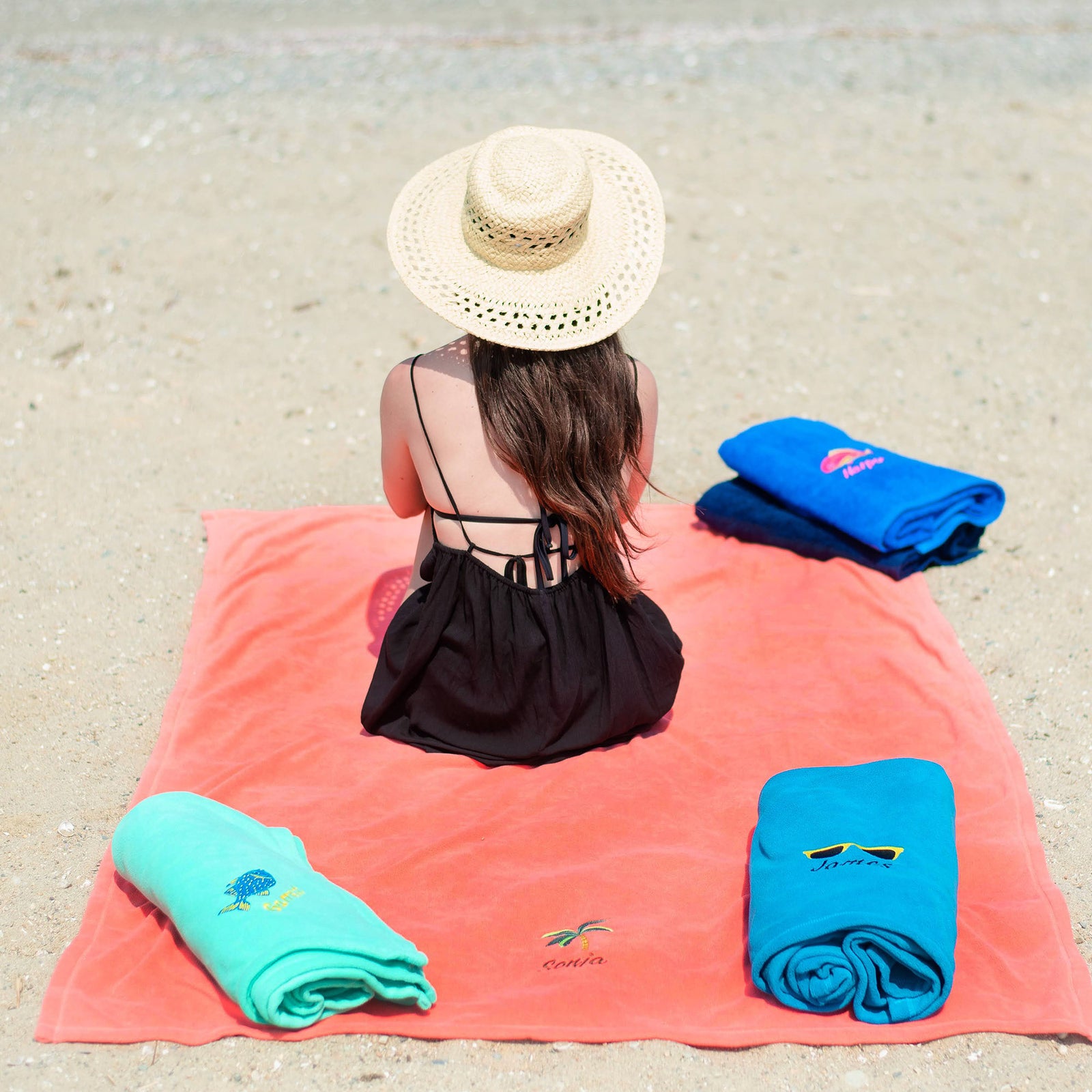 American Blanket Company - Peaceful Touch Beach Blankets - Pictured is a young woman in a beach hat sitting on a coral  peaceful touch beach blanket with a palm tree embroidery along with two other peaceful touch beach blankets and a pair of cotton beach towels.